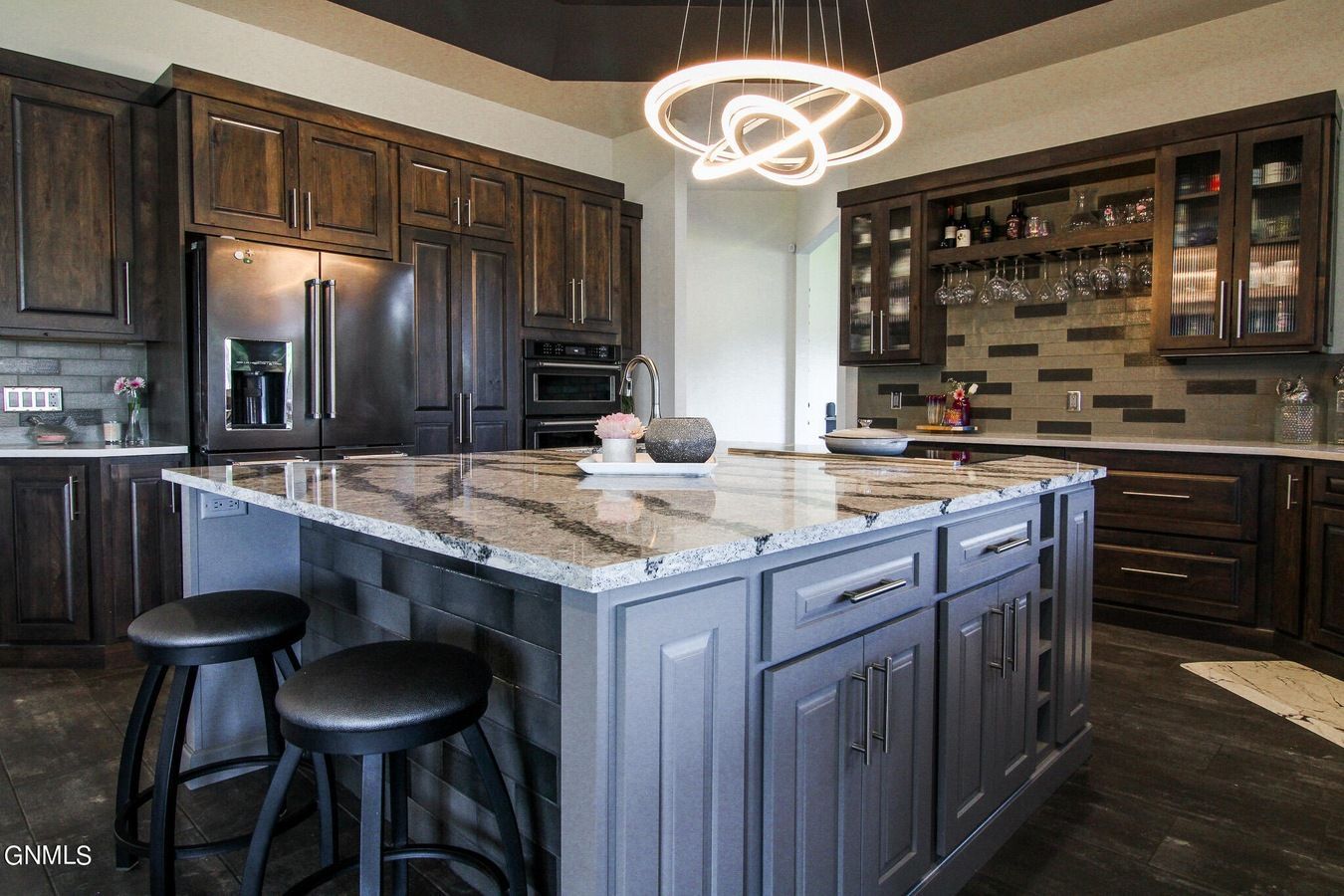 A modern kitchen with dark wooden cabinets, a large marble island, two stools, and a unique circular chandelier.