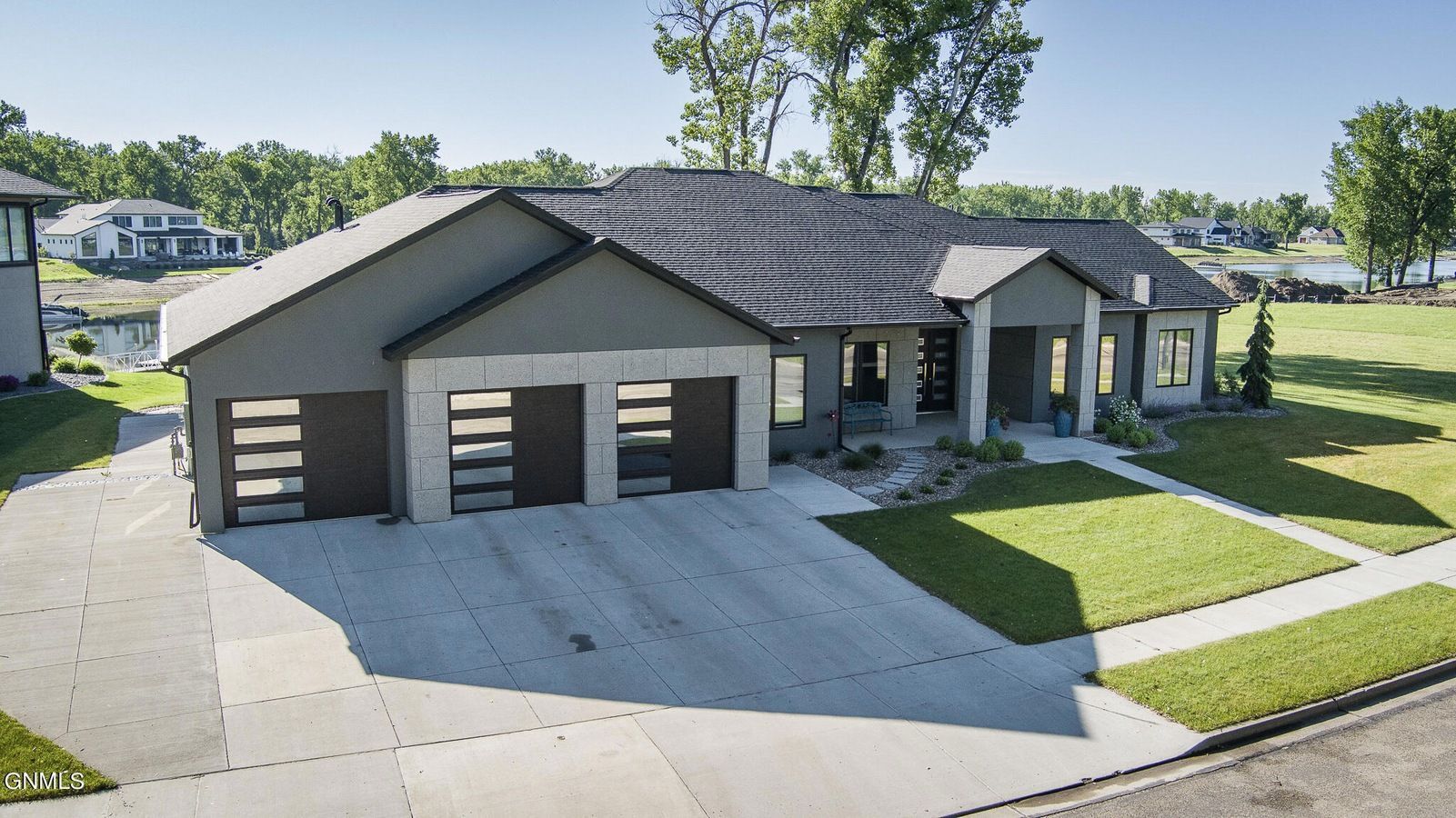 A modern, grey suburban house with a three-car garage, stone accents, and a paved driveway under a clear blue sky.