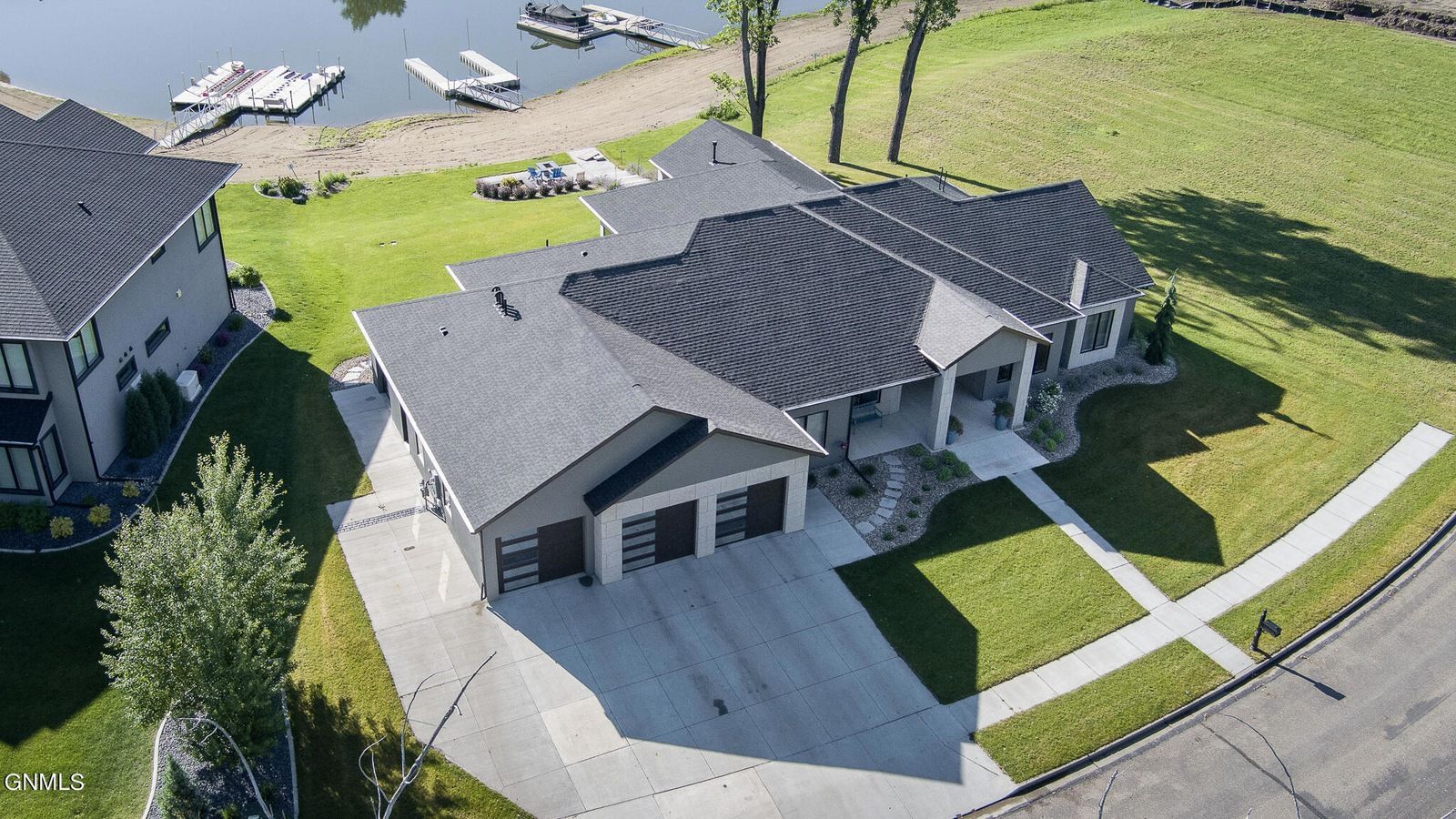 An aerial view of a single-story house with a three-car garage and dark shingled roof, set near a waterfront.