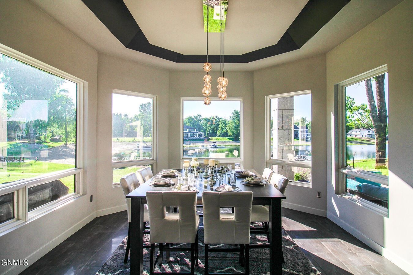 A formal dining room featuring a dark table with chairs set for six, surrounded by large windows overlooking a lake.
