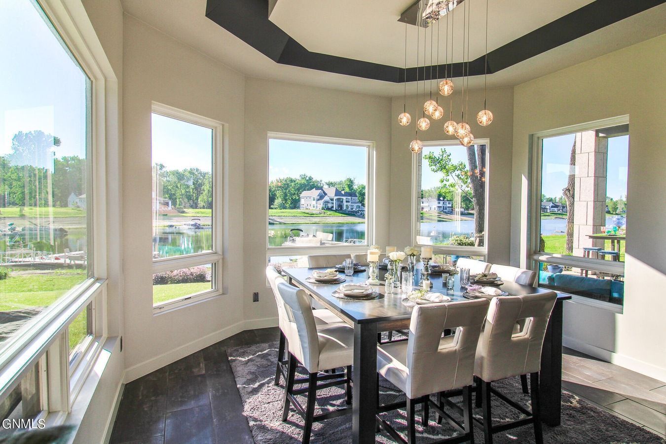 A bright dining room with floor-to-ceiling windows overlooking a lake, featuring a modern table, chairs, and chandelier.