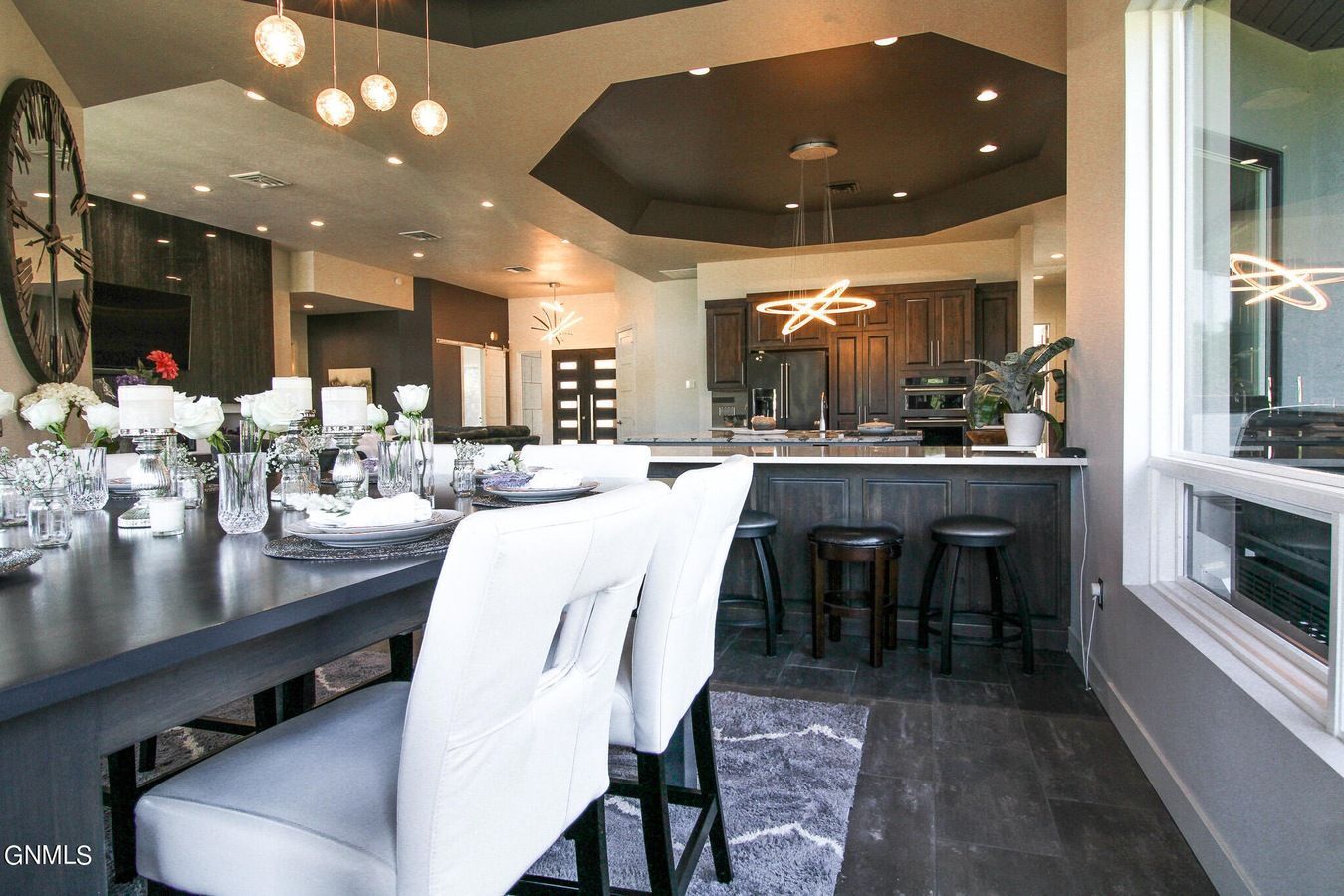 A dining room with a long table and white chairs, opening into a dark-toned kitchen with bar stools and pendant lighting.