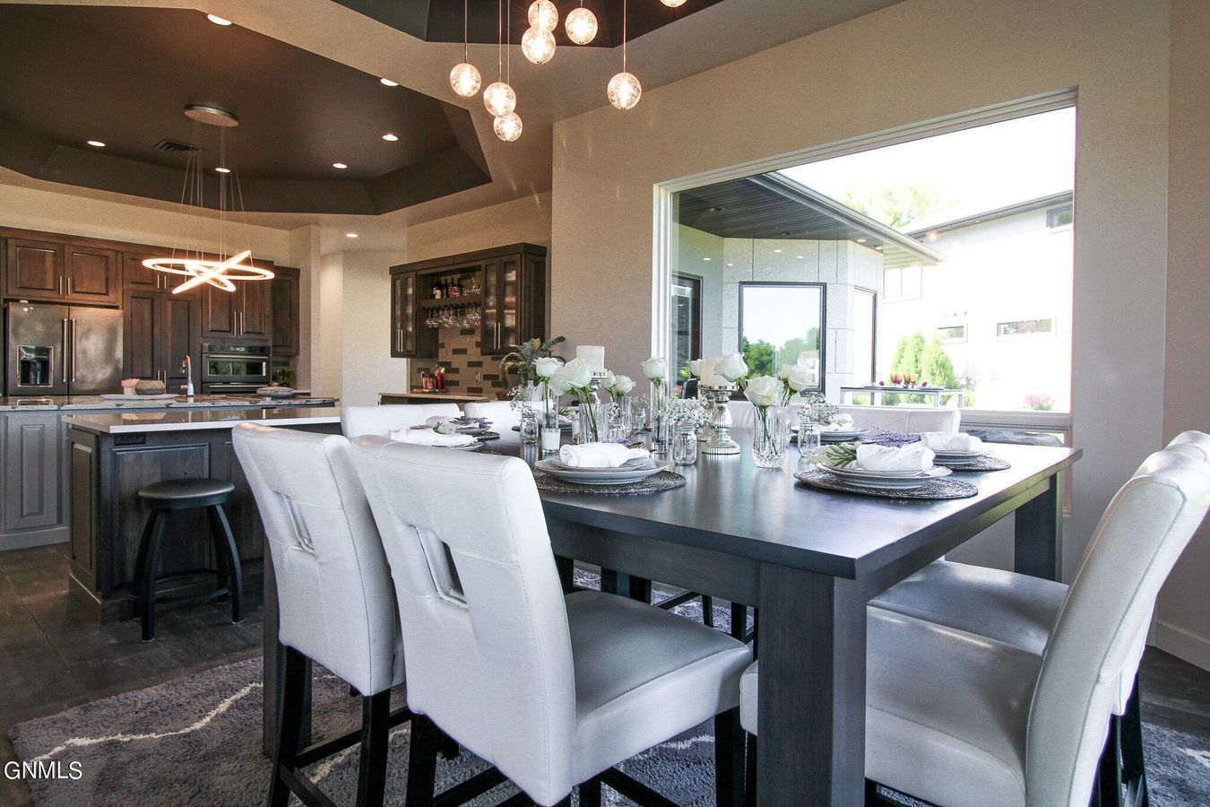 A modern dining room with a dark wooden table, upholstered chairs, and a kitchen with dark cabinets in the background.