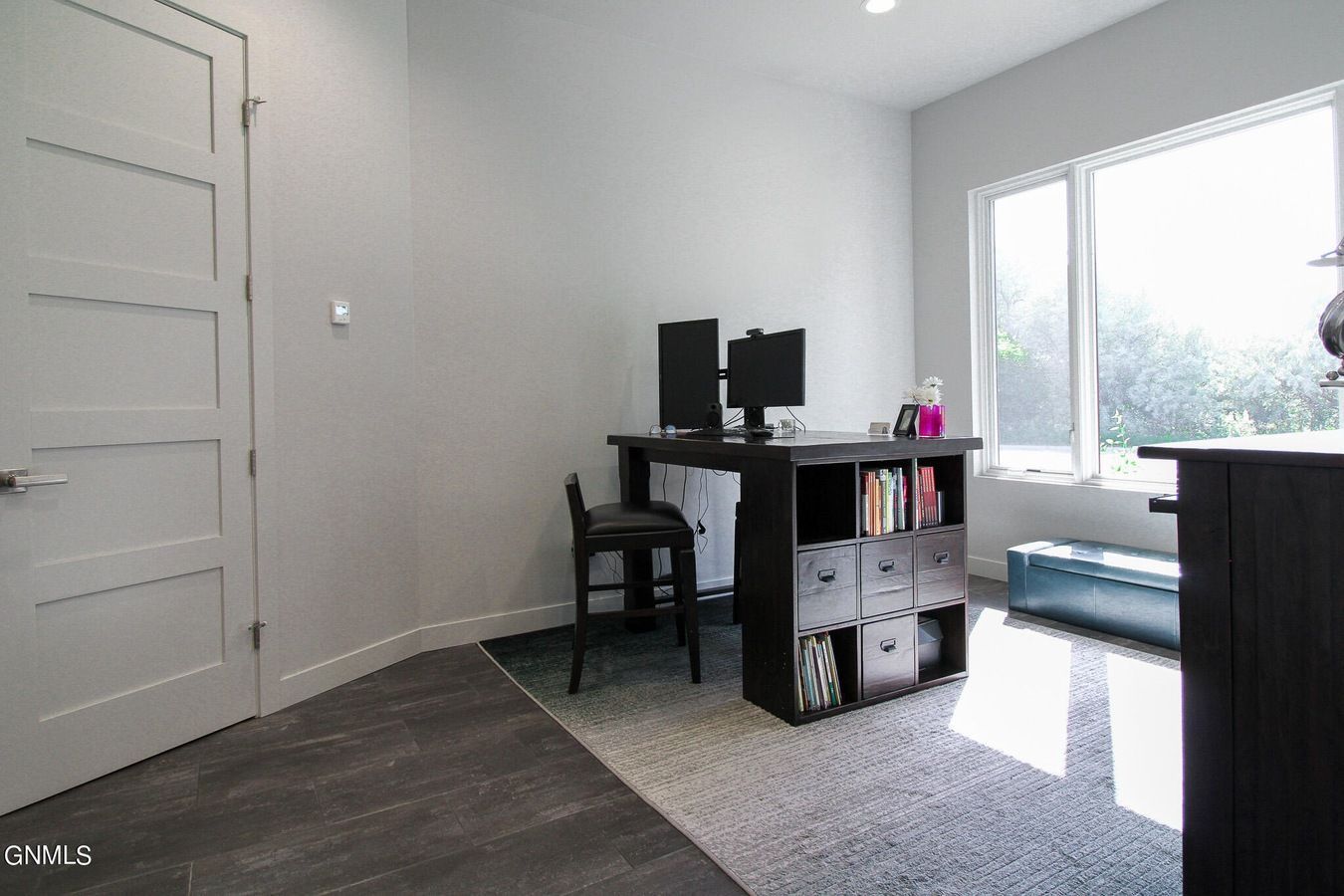 A home office featuring a desk with two computer monitors and storage cubbies on a patterned rug near a large window.