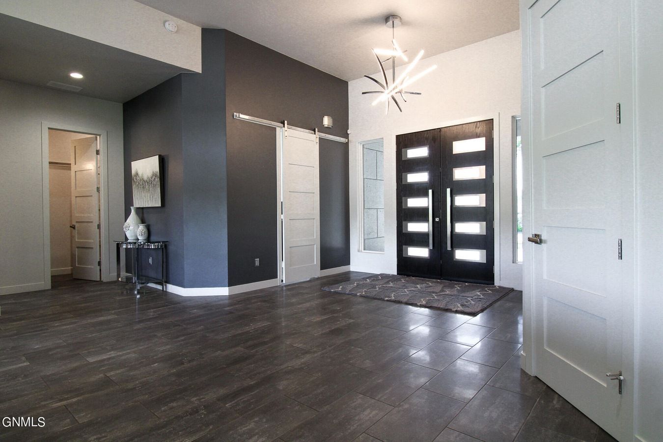 A modern foyer with dark grey walls, wood-look tile flooring, a geometric chandelier, and double front entry doors.