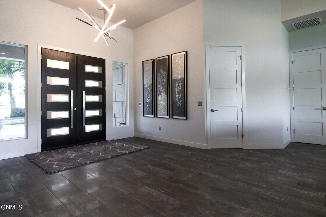 A bright entryway with dark tile floors, a modern double door, a geometric light fixture, and three framed wall prints.