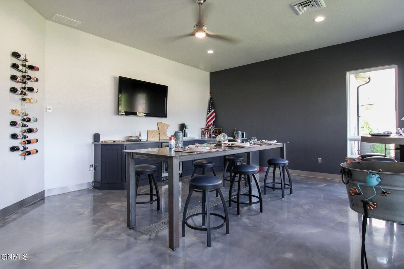 A contemporary entertainment room with a bar-height table, black stools, a mounted TV, wall-mounted wine rack, and flag.