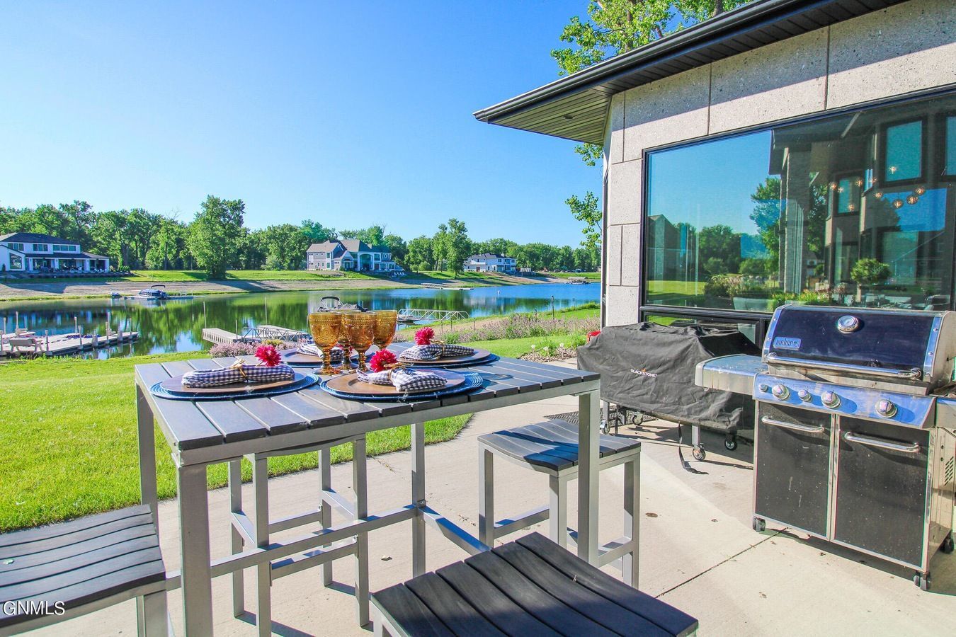A patio with a dining set and grill overlooking a lake and houses under a clear blue sky.