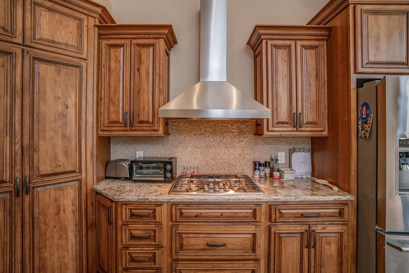 A kitchen stove area featuring brown wood cabinetry, a stainless steel range hood, and a light-colored stone countertop.