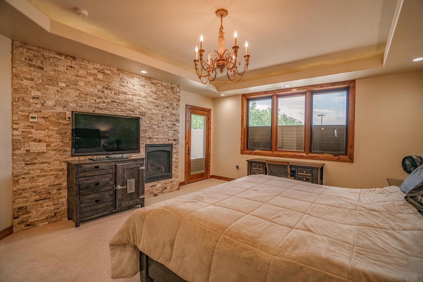 A master bedroom with a stone feature wall, a television, a fireplace, a chandelier, and a bed with a tan comforter.