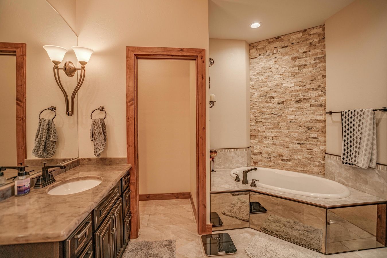 A bathroom featuring a stone wall, a soaking tub with mirrored accents, a vanity with a stone countertop, and a doorway.