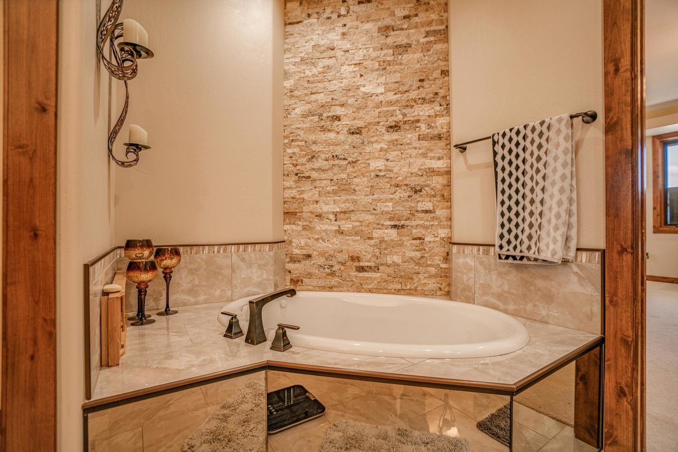 A corner bathtub with a tiled deck and a stone accent wall, featuring a nearby wall sconce and a towel rack.