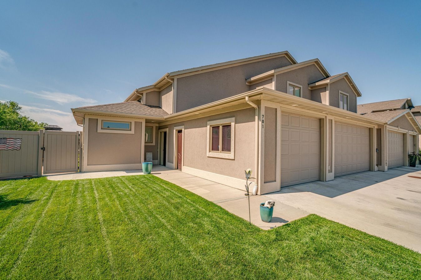 A tan, two-story townhome with attached garages, a concrete driveway, and a vibrant green lawn under a clear blue sky.