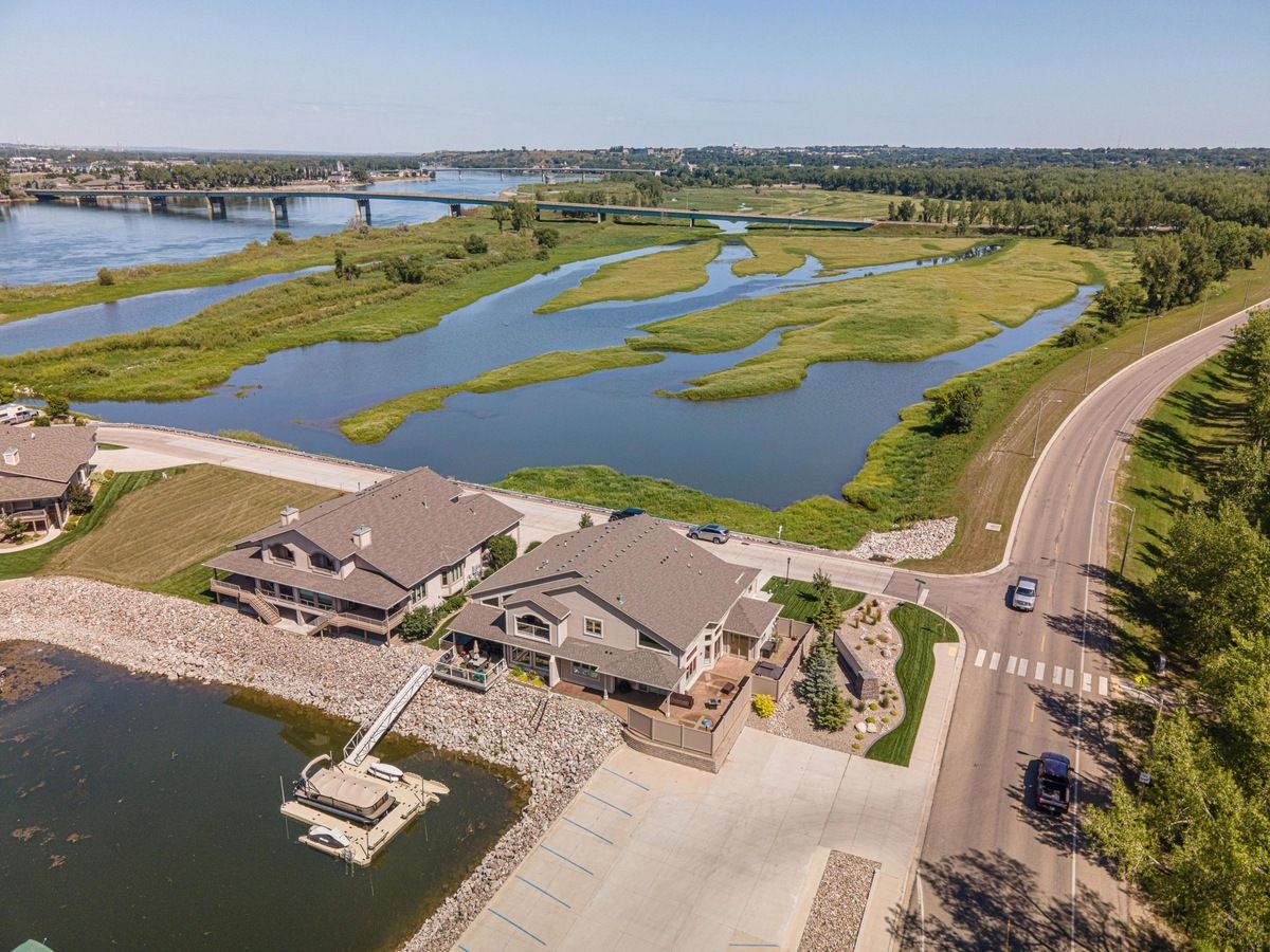 An aerial view of two residential homes along a river, with a dock, a nearby bridge, and lush wetlands in the background.