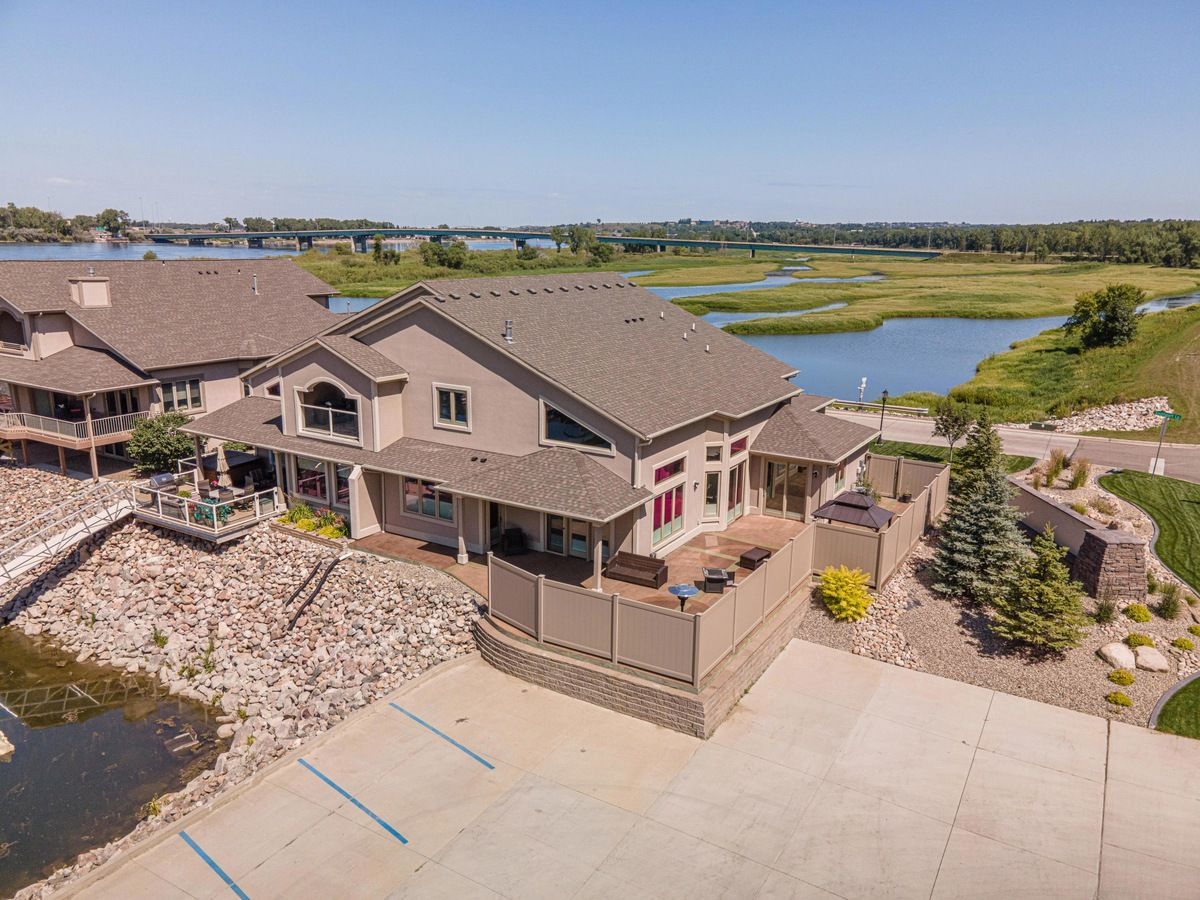 Aerial view of a beige multi-story house near a wetland, with a paved parking area and a bridge in the distance.