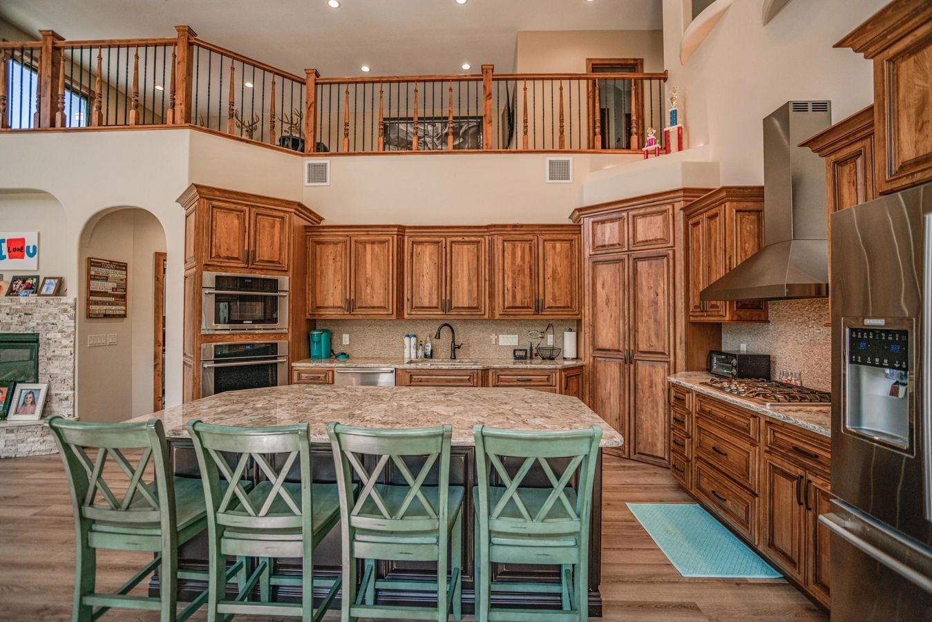 A spacious kitchen featuring wood cabinets, granite countertops, a large island with four green chairs, and a loft area.