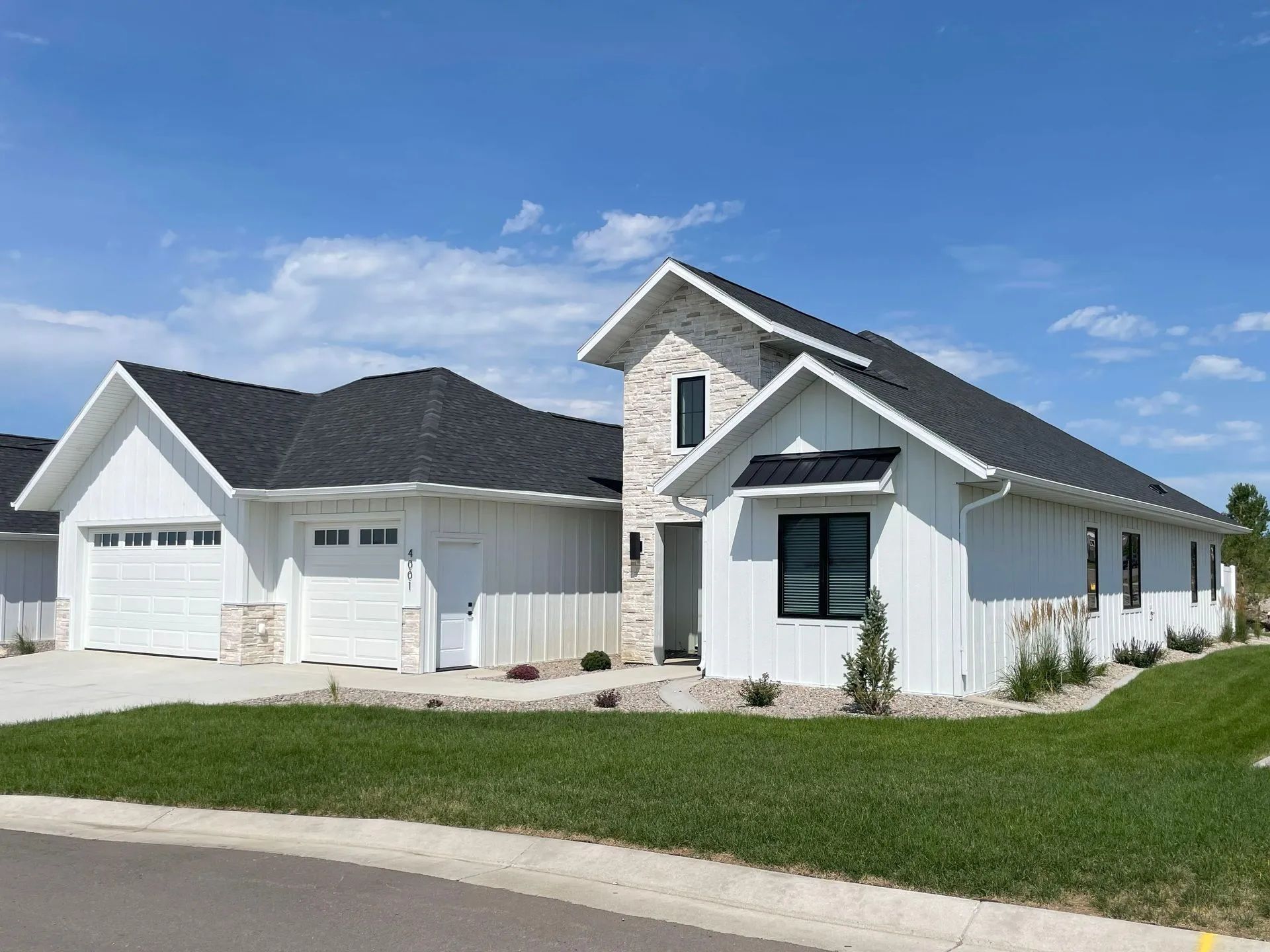 A modern one-story suburban house with white vertical siding, stone accents, and a dark shingled roof under a blue sky.