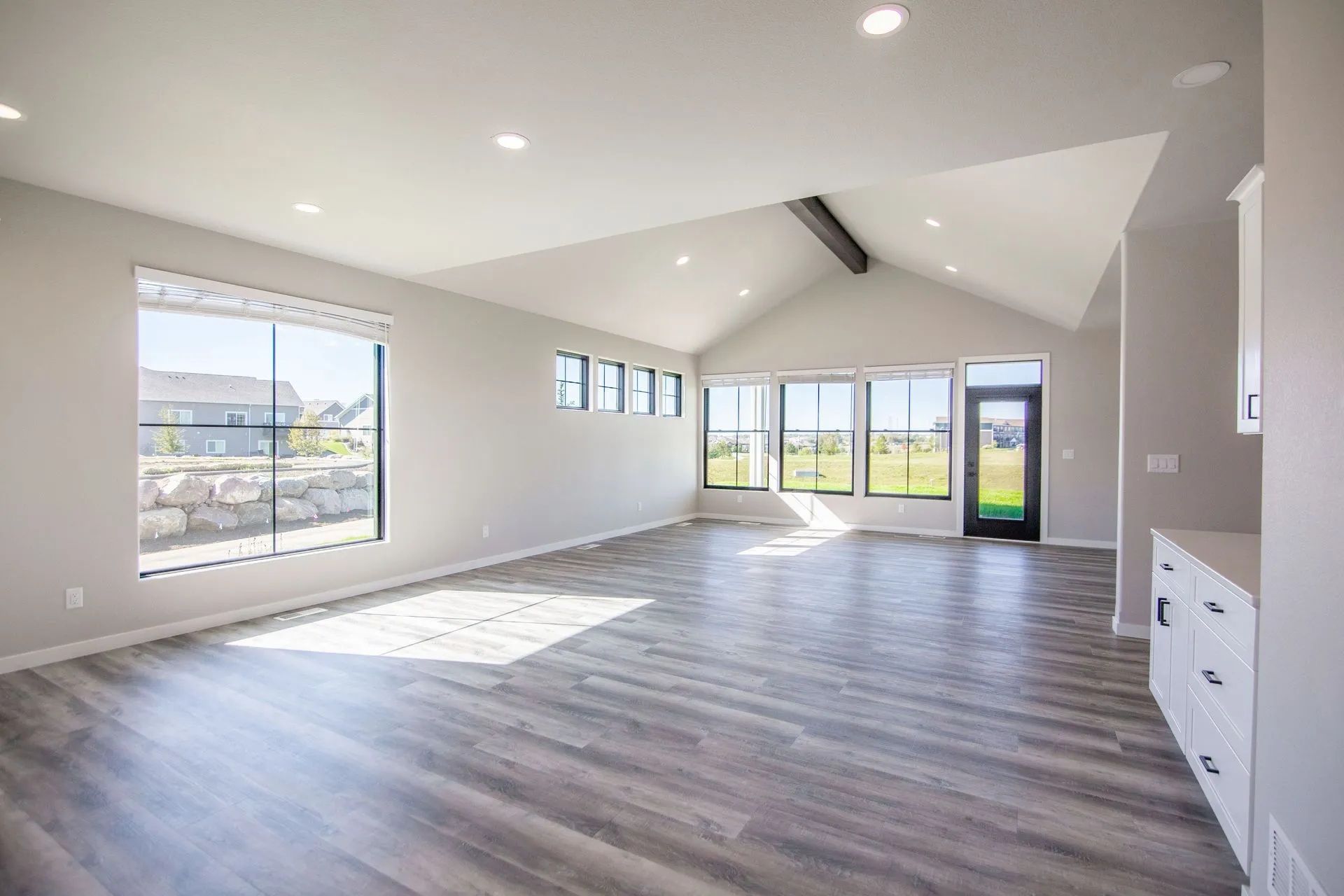 A bright, empty living room with light gray walls, wood-look flooring, a vaulted ceiling, and large windows.
