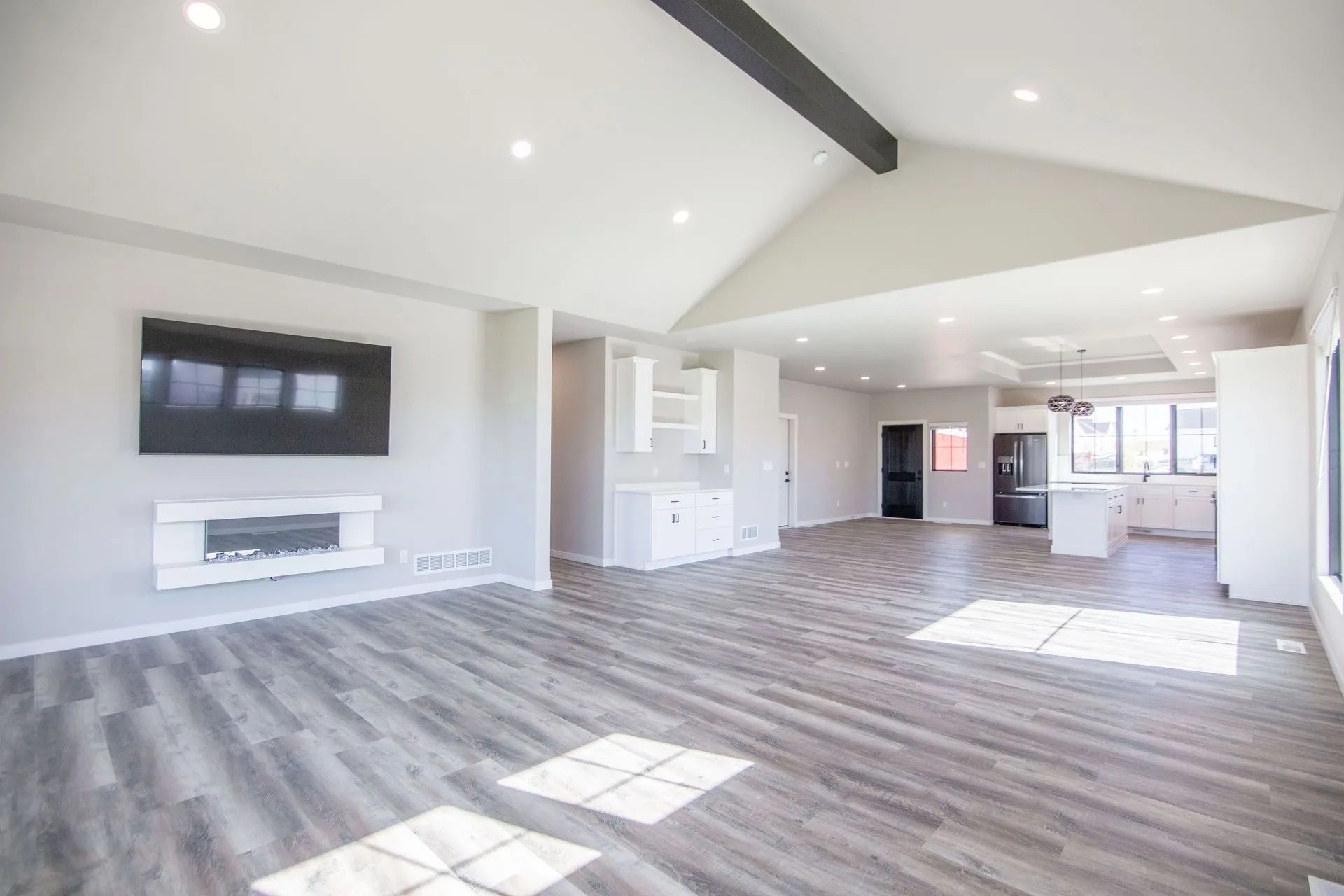 An open-concept living area with light grey wood floors, vaulted ceilings, a wall-mounted TV, fireplace, and kitchen.