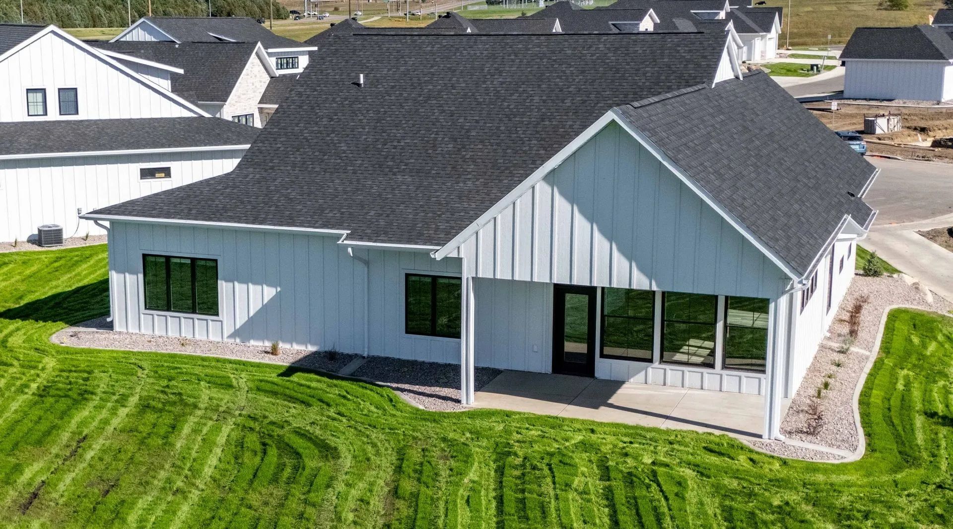 A modern white farmhouse with a black roof and a rear covered patio sits on a green, freshly mowed lawn.