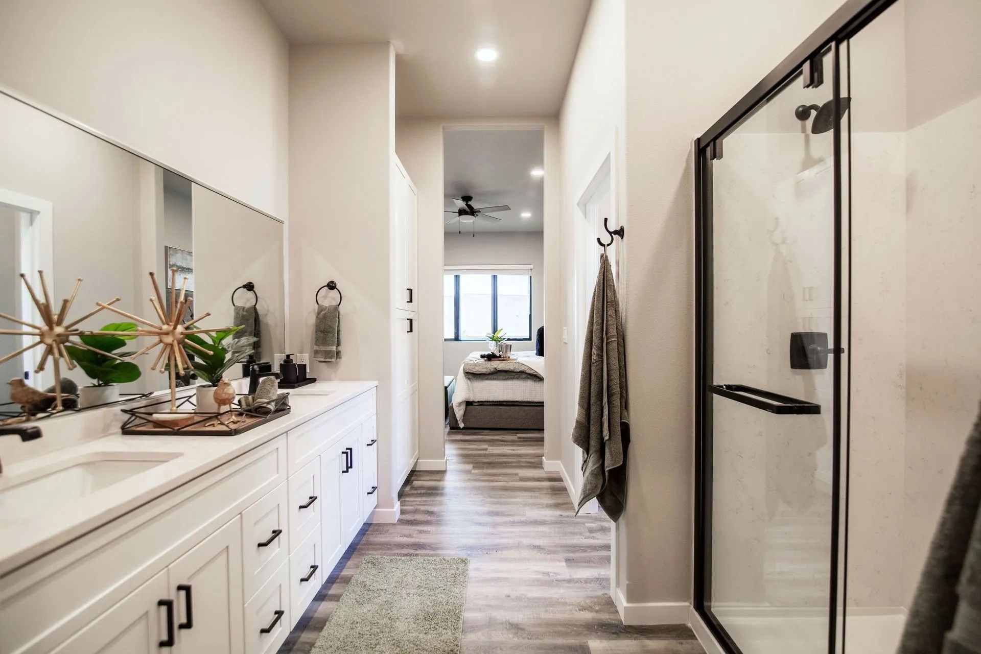 A modern bathroom with a white vanity, a long mirror, and a glass-enclosed shower, with a view of a bedroom down the hall.