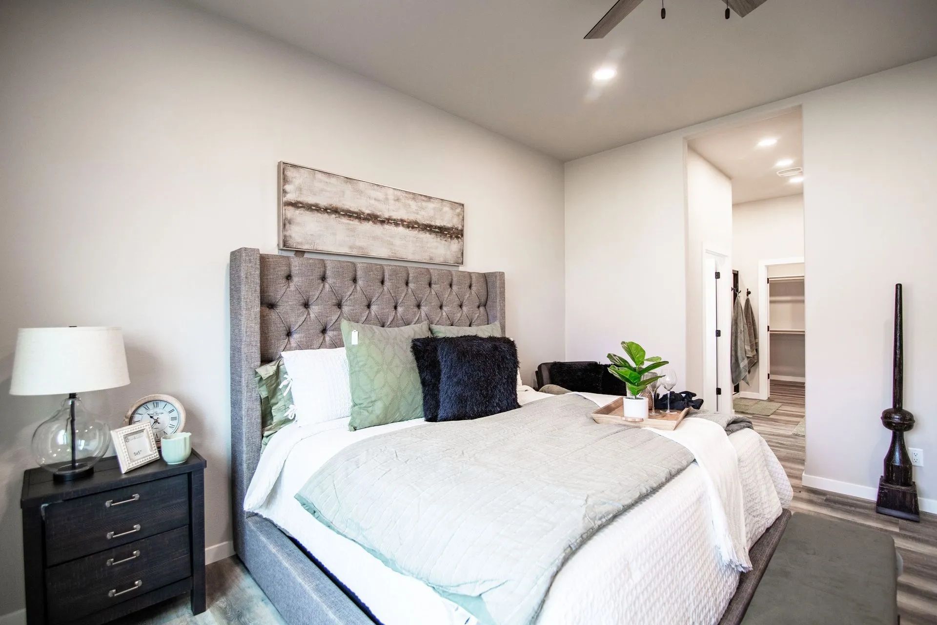 A bedroom featuring a gray tufted headboard, white bedding, green pillows, a dark nightstand, and a view into a closet.