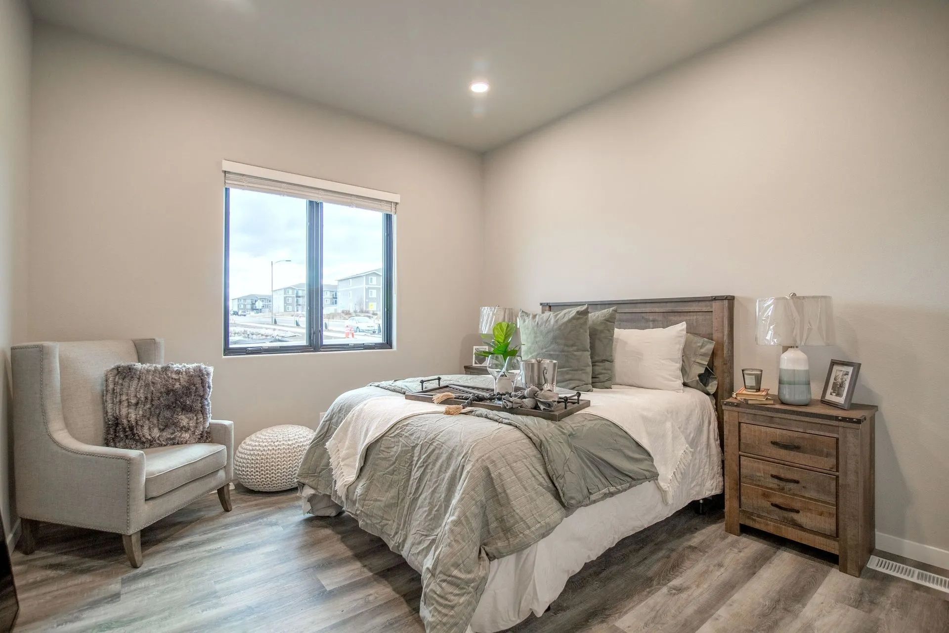 A modern bedroom features a gray armchair, a queen bed with neutral bedding, and a rustic wooden nightstand.