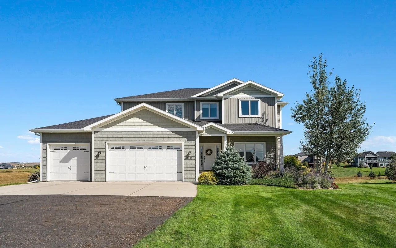 A multi-level suburban home with grey siding, a three-car garage, and a manicured lawn under a clear blue sky.