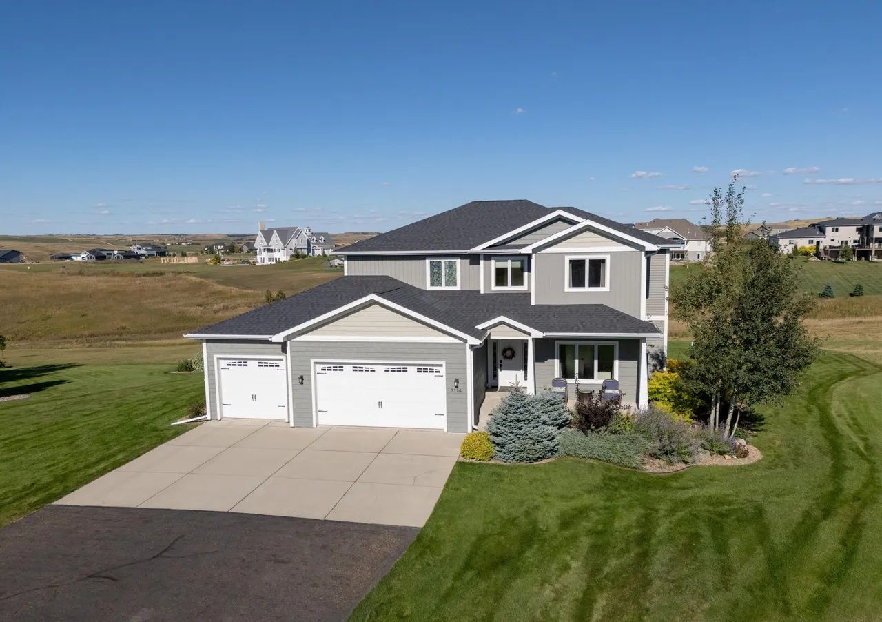 A two-story grey suburban house with a three-car garage and asphalt driveway, surrounded by green lawns and open fields.