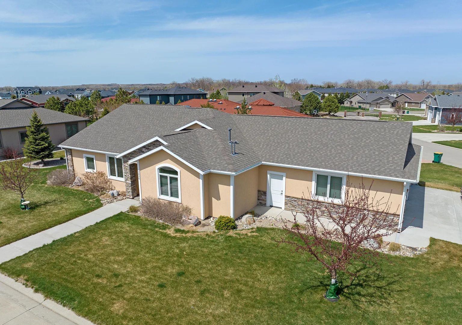 An aerial view of a tan-colored single-story suburban house with a gray shingle roof on a sunny day.