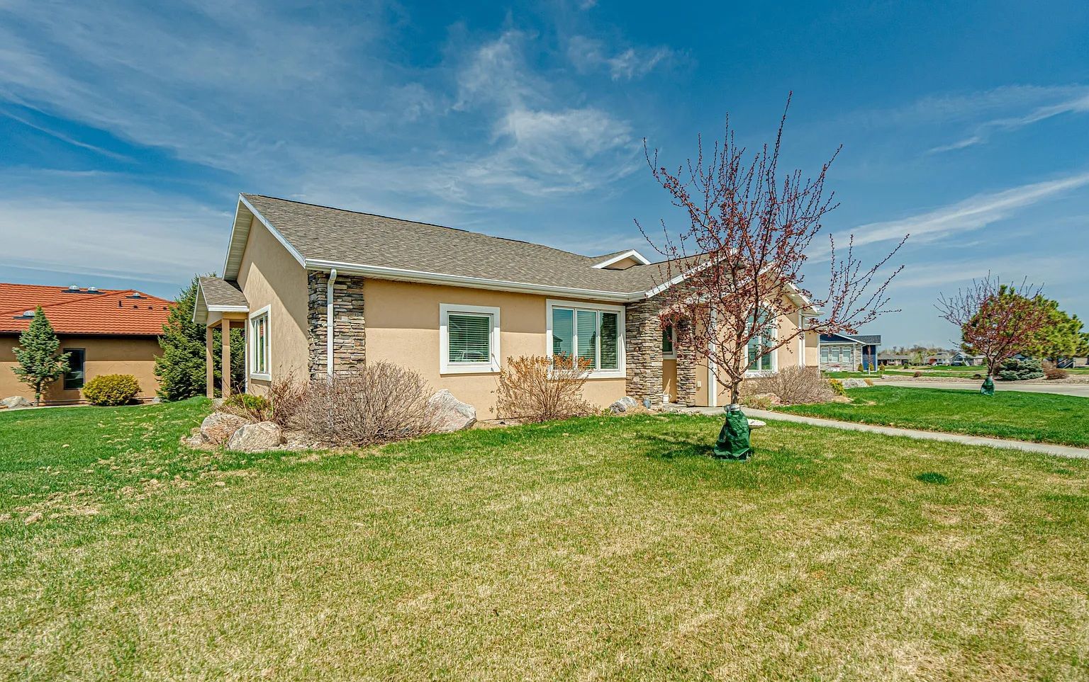 A beige, single-story house with a stone facade and shingled roof sits on a sunny, grassy lot under a blue sky.