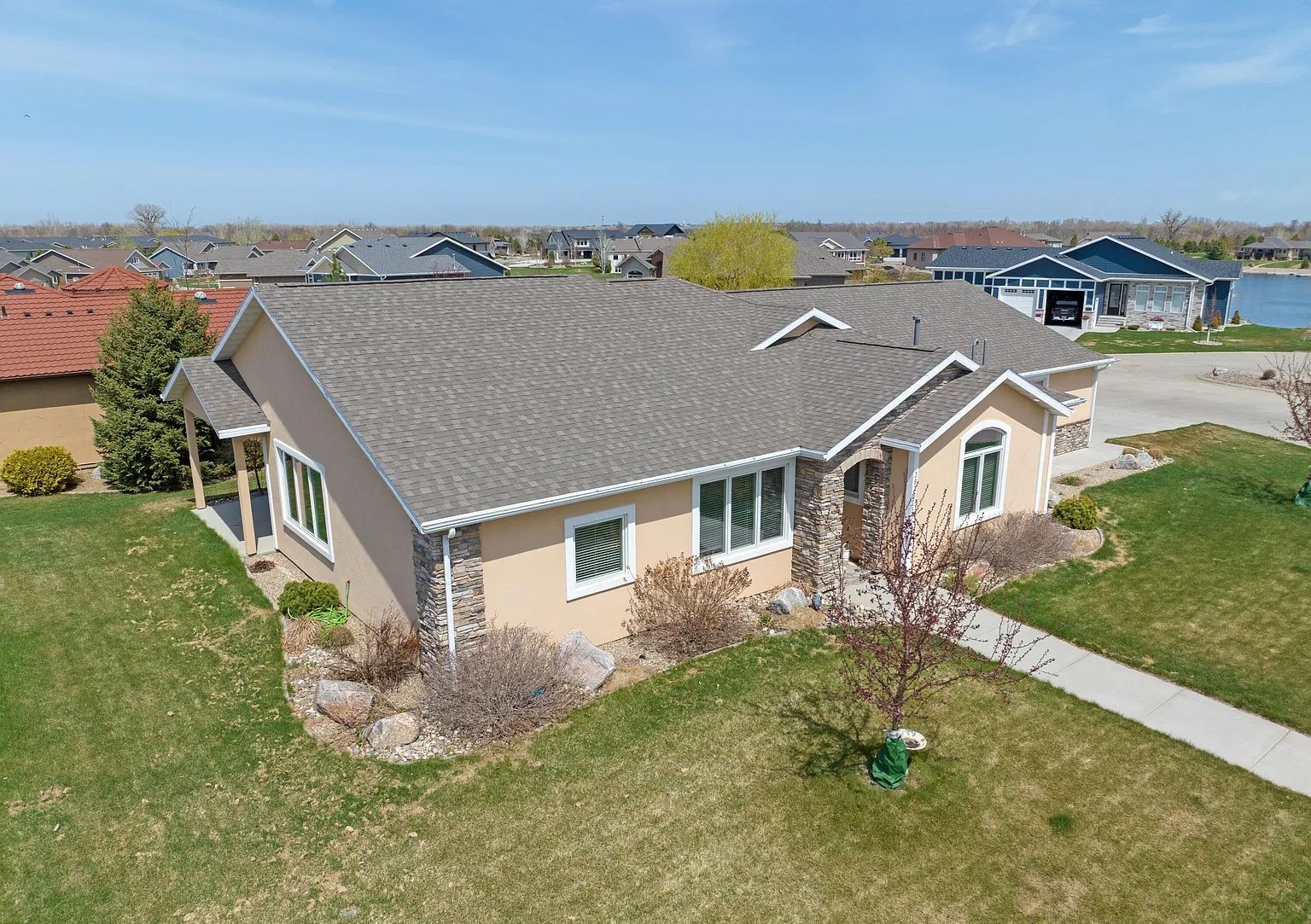 An aerial view of a tan single-story suburban house with a gray roof, stone accents, and green lawn on a sunny day.