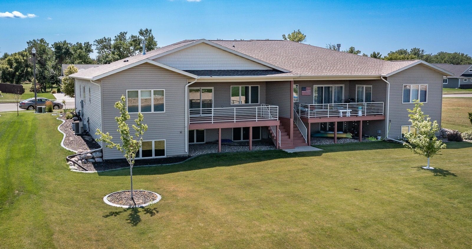 A light-gray, two-story house with a wooden deck, a light-tiled roof, and a grassy yard under a clear blue sky.