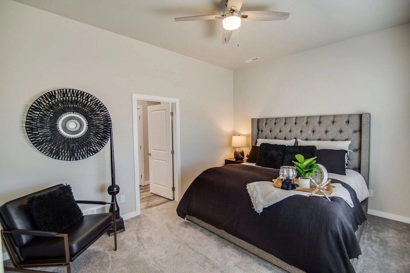 A bedroom with a tufted gray bed, a black armchair with a decorative wall piece, and a doorway, all on gray carpet.