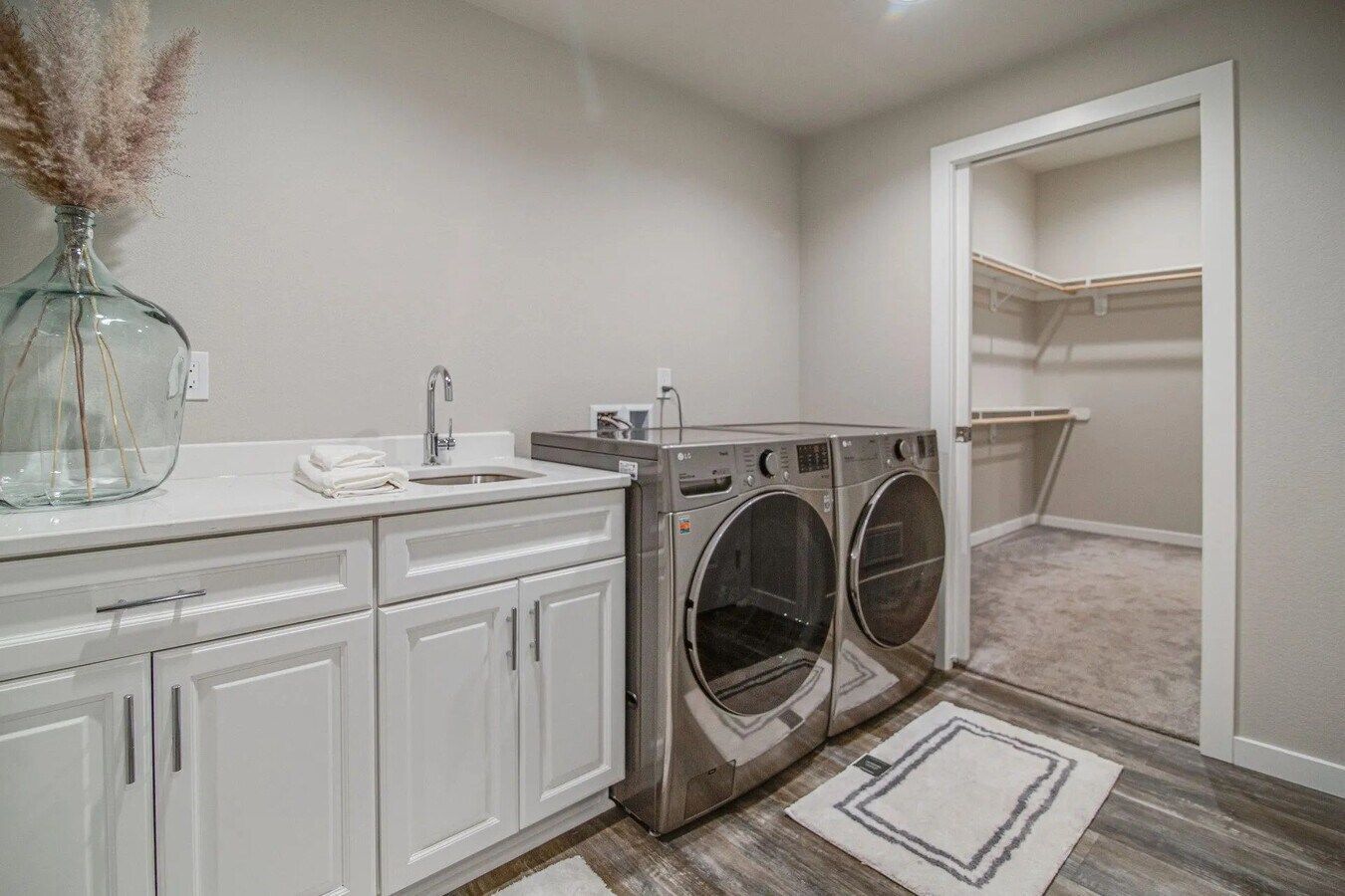 A modern laundry room featuring white cabinetry, a sink, stainless steel washer and dryer, and an open walk-in closet.