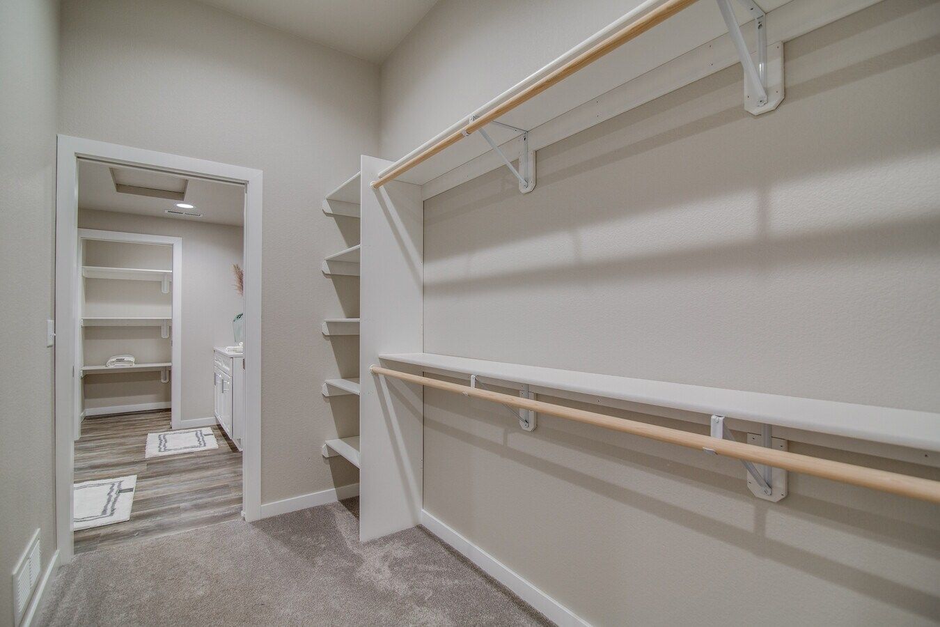 A walk-in closet featuring white wooden shelving and hanging rods with carpeted floors and a view into a bathroom.