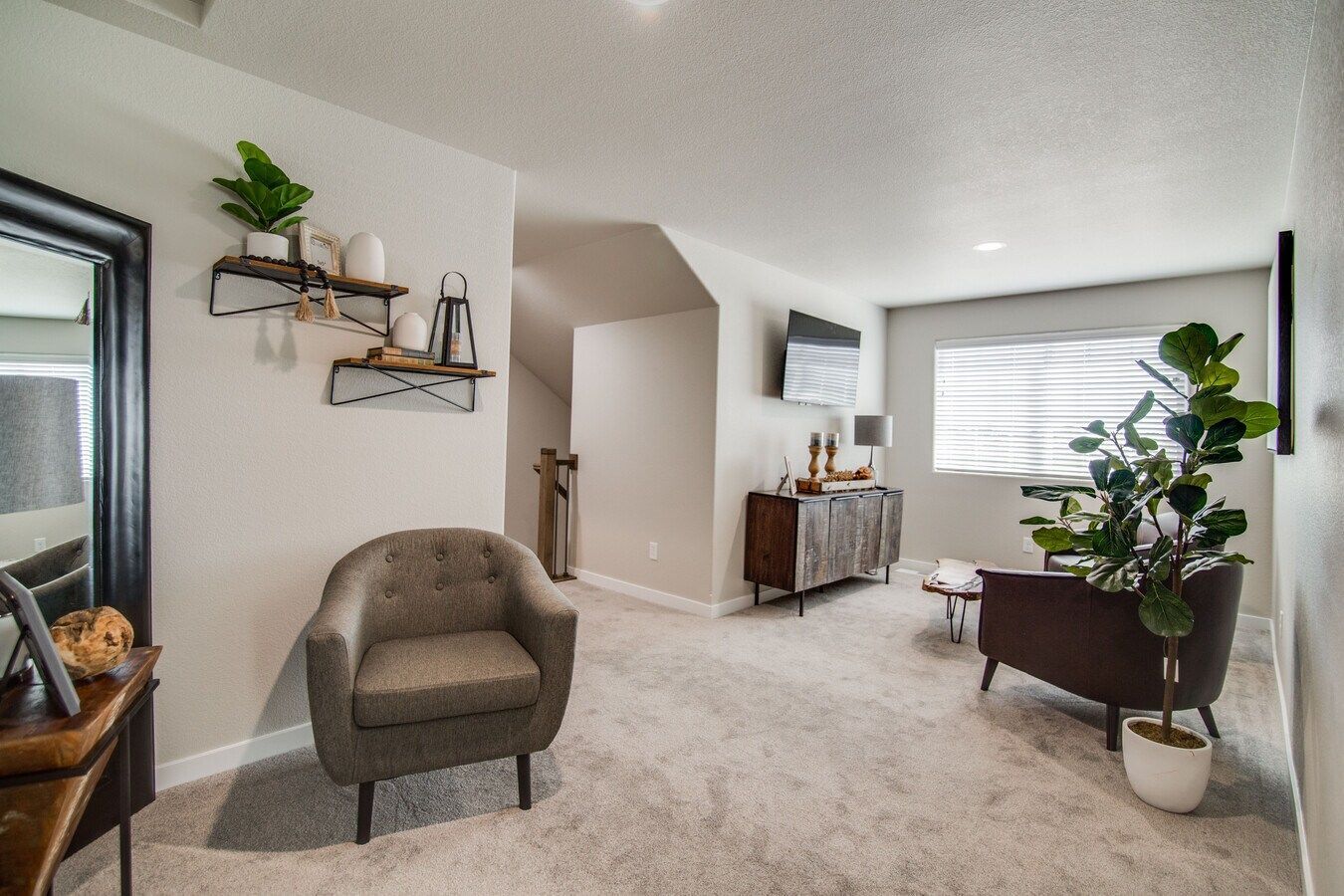 A neutral-toned living room featuring a gray armchair, a potted fiddle leaf fig, wall shelves, and a mirror.