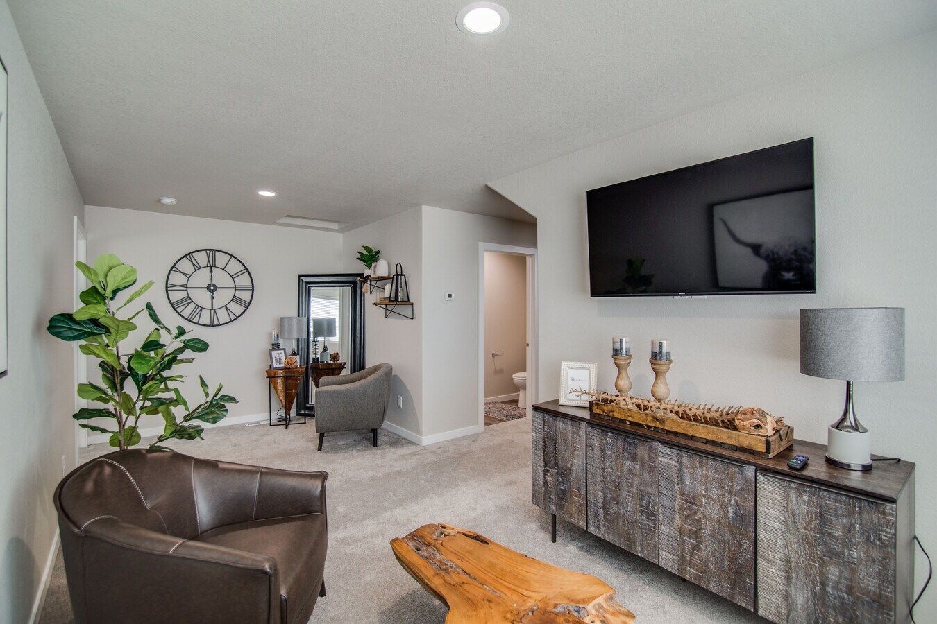 A living room with light walls, gray carpet, a leather armchair, a wood coffee table, and a media console with a wall TV.