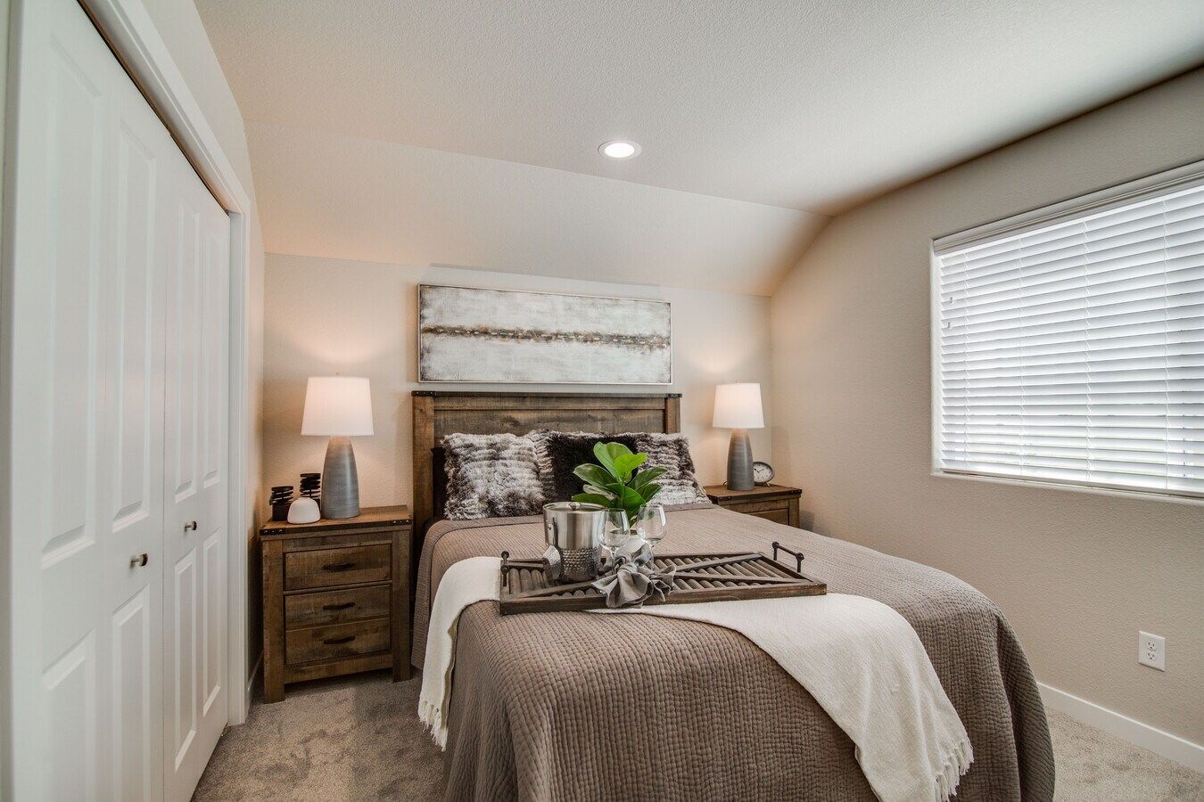 A bedroom with a queen bed, wooden nightstands with lamps, and a window with blinds, decorated in neutral, earthy tones.