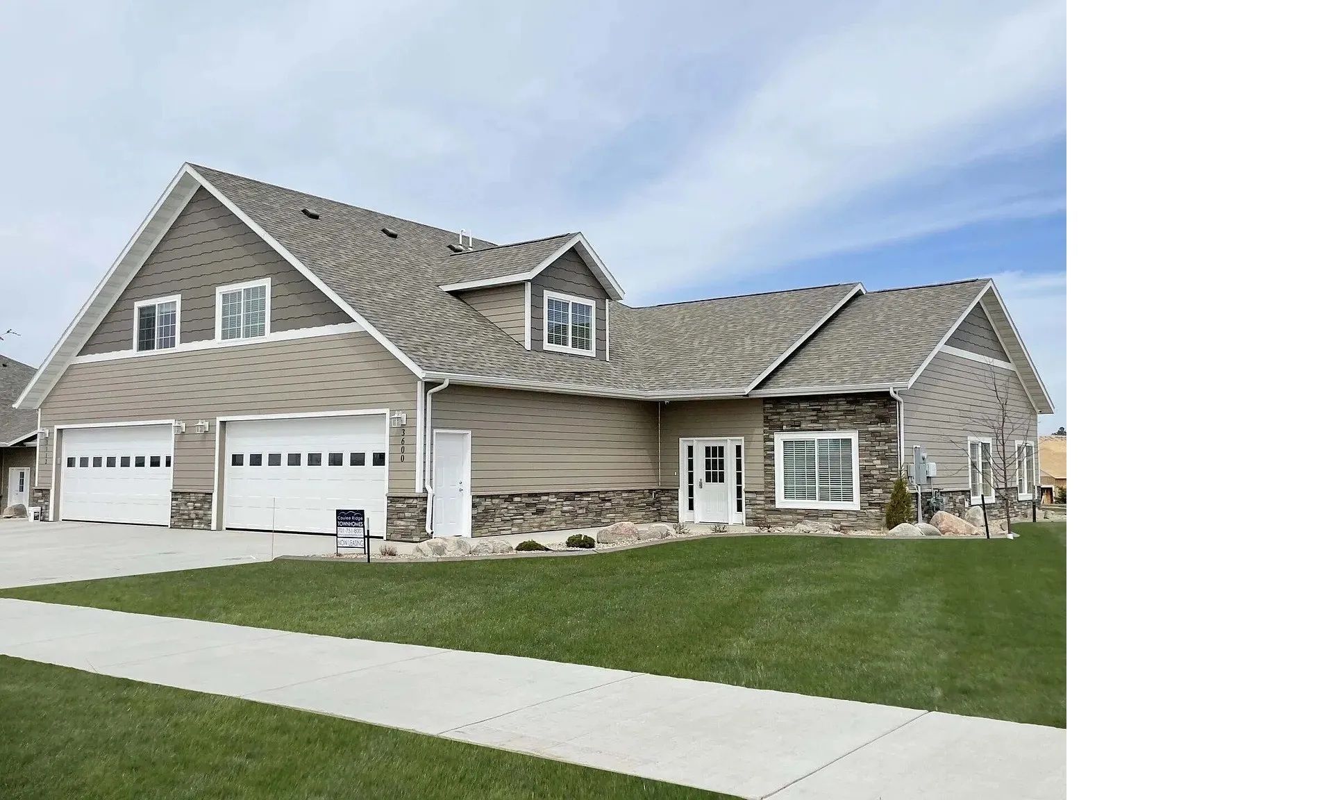 A tan, two-story house with a white two-car garage, stone accents, and a paved walkway in front under a blue sky.