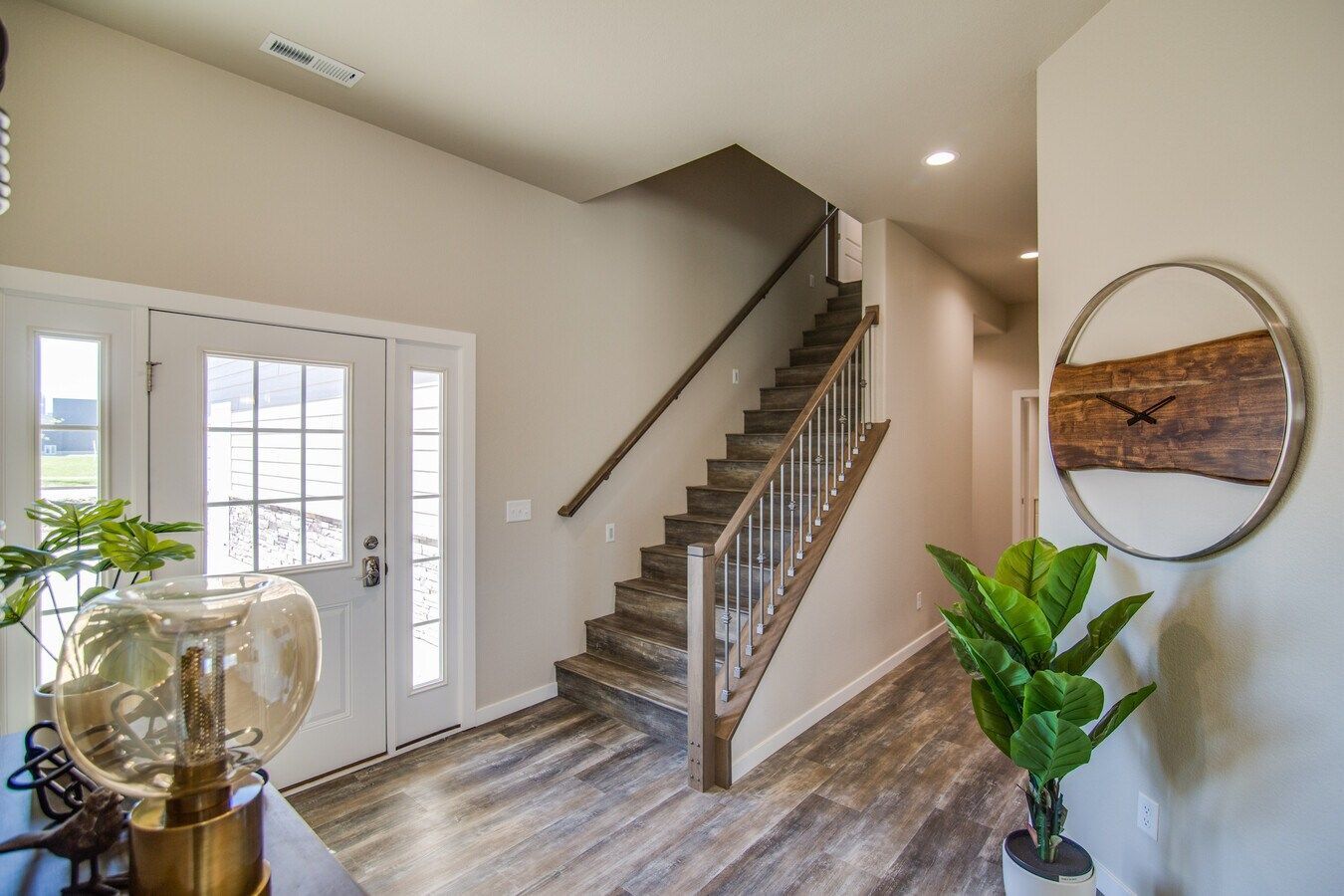 A bright entryway features wood-look floors, a white front door, a staircase with a wooden railing, and a wall clock.