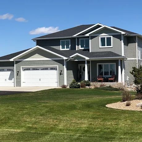 A two-story, gray house with white trim, a three-car garage, and a lush green lawn under a clear blue sky.