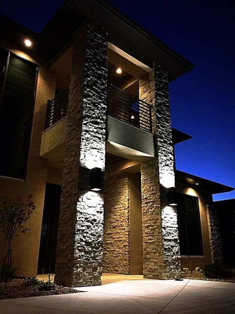 Exterior view of a modern stone house at night, illuminated by warm wall-mounted lights on two large stone pillars.
