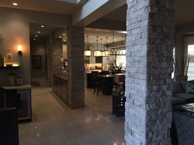 An interior view of an open-plan home with light-colored tile floors, stone-clad pillars, and a kitchen in the background.