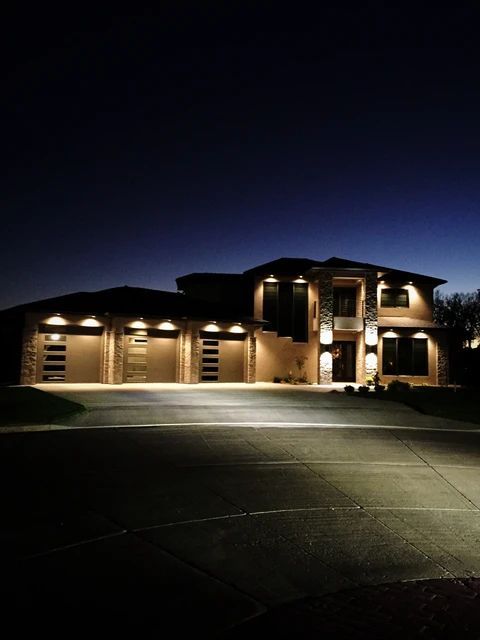 A modern two-story house with a three-car garage is illuminated by exterior lighting at dusk under a dark blue sky.
