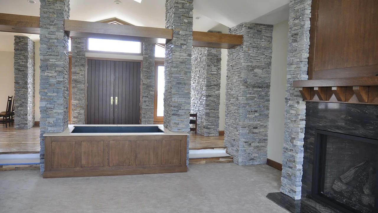 A room with a stone fireplace, large stone columns, wooden beams, a wooden accent wall, and a low wooden cabinet.