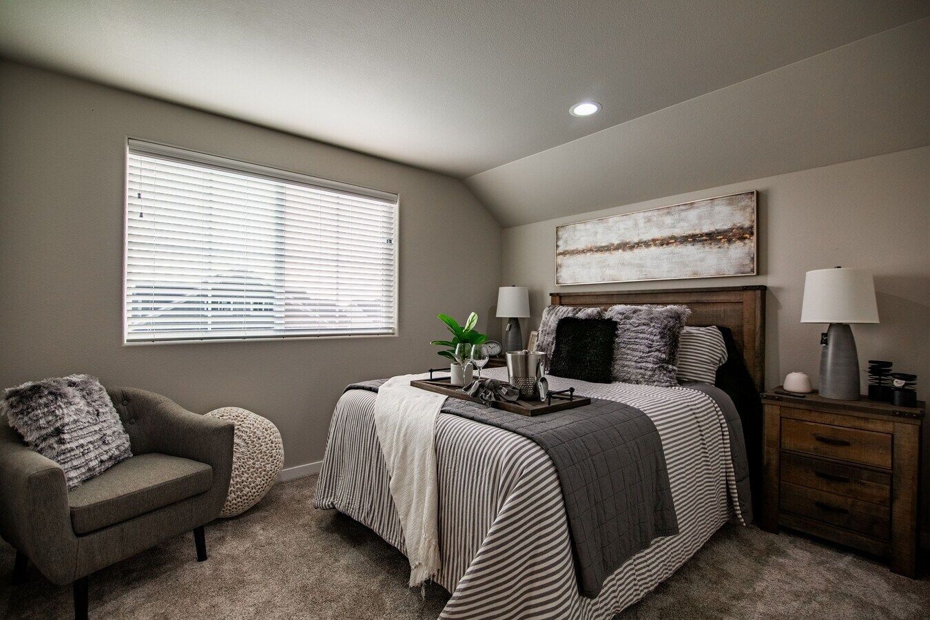 A cozy bedroom with a patterned bedspread, dark wood furniture, a gray armchair, and a window with white blinds.