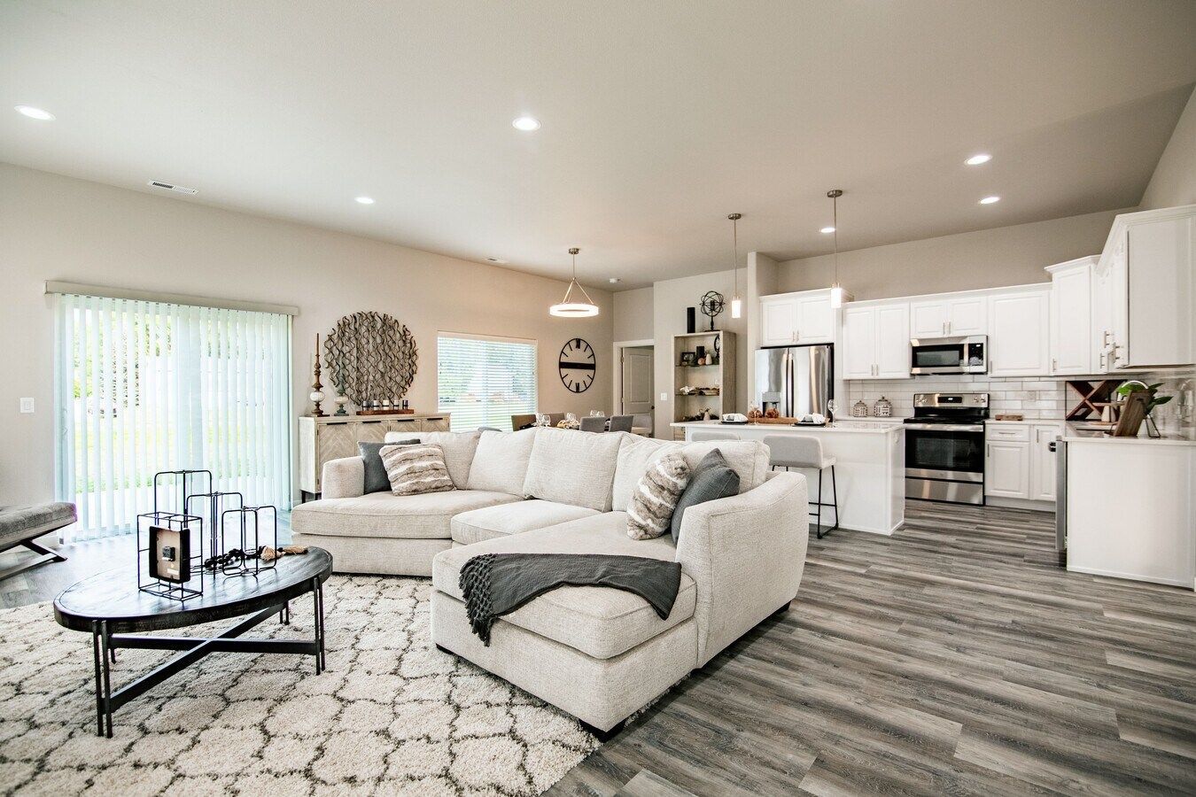 An open-concept living room with a white sectional sofa, patterned rug, glass coffee table, and white modern kitchen.