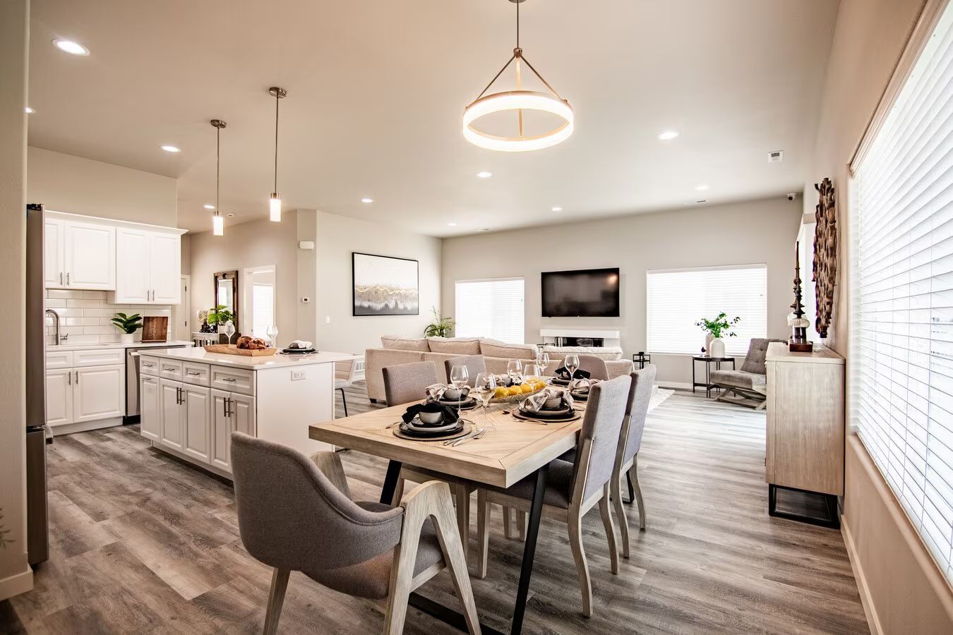 Open-concept kitchen, dining, and living area with white cabinets, a light wood dining table, and gray vinyl plank floors.