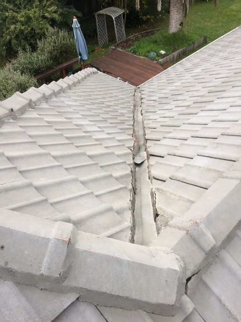 Overhead view of a light gray tiled roof showing a central valley with visible cracks and separation in the mortar, with a backyard deck and garden visible below. Overhead view of a light gray tiled roof showing a central valley with visible cracks and separation in the mortar, with a backyard deck and garden visible below.